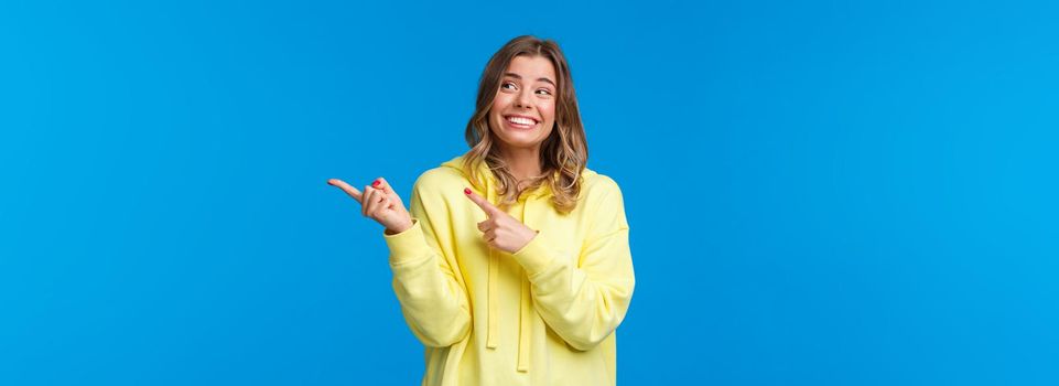 Cheerful hipster girl in yellow hoodie, with blond short haircut, pointing and looking right side with pleased happy smile, promote product or company services, stand blue background.