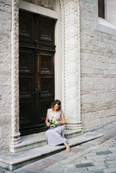 Bride with flowers in her hands sits on the steps near the wooden carved door. High quality photo