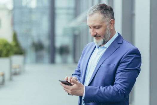 Senior handsome gray-haired man in a suit stands on the street, holds a phone in his hand, dials a number, calls someone, smiles.