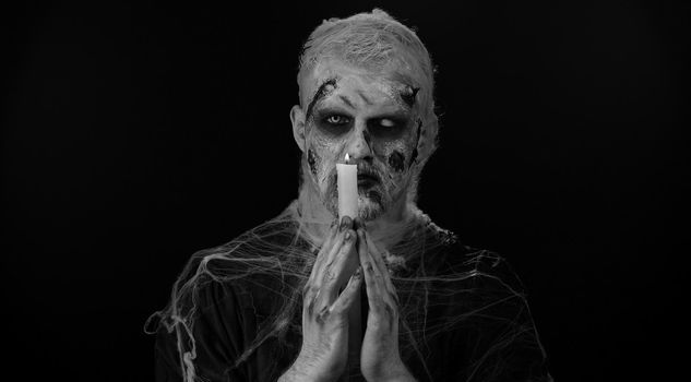 Frightening man with Halloween zombie bloody wounded makeup spells conjures over a candle. Horror theme. Sinister undead guy isolated on studio black background. Voodoo rituals. Fashion body art