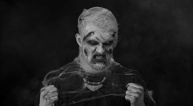 Frightening man with Halloween zombie bloody wounded makeup, trying to scare, face expressions. Screaming. Sinister undead guy isolated on studio black background. Voodoo rituals. Fashion body art