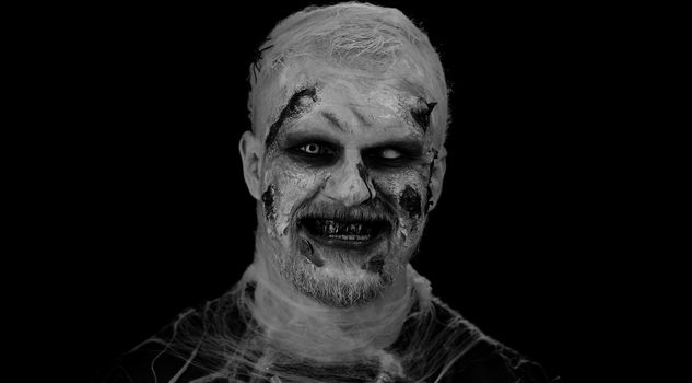Scary man with Halloween zombie bloody wounded makeup, trying to scare, showing tongue. Convulsions. Sinister undead guy isolated on studio black background. Voodoo rituals. Fashion body art