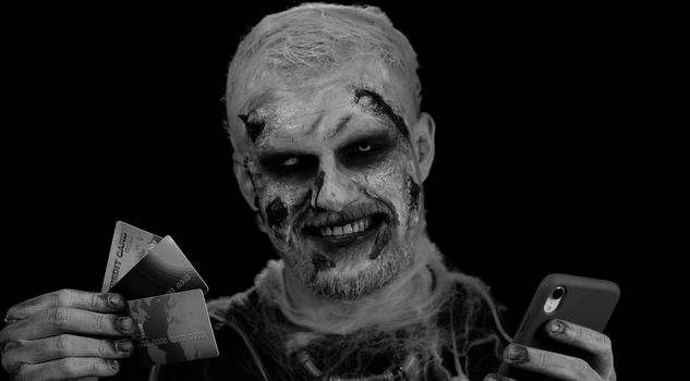 Sinister man with horrible scary Halloween zombie make-up using credit bank cards and mobile phone, transferring money purchases online shopping. Dead guy with wounded bloody scars face, black room