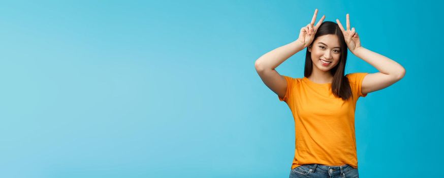 Cute tender asian girlfriend act silly lovely smile, tilt head show playfully ears hold peace victory signs on head, grinning toothy, express joy happiness and delight, stand blue background.