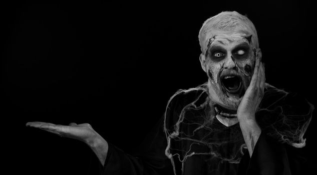 Wow surprise. Zombie man with fake wounds scars make-up showing thumbs up and pointing empty place, advertising area for commercial text logo, copy-space for goods promotion. Halloween ugly dead guy