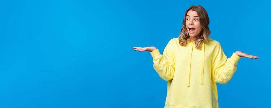 Girl weighing two choices as making decision, look away picking between products she holding in arms on left and right side, standing excited over blue background.