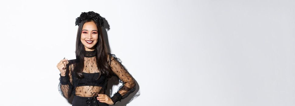 Image of beautiful asian woman in halloween costume, showing credit card and smiling, standing in gothic lace dress over white background.