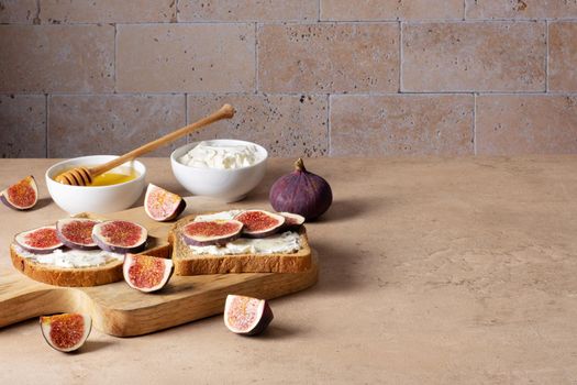 Bruschetta with figs, cream cheese and honey lies on wooden cutting board on light beige table against brick wall. Ingredients. Healthy food. Selective focus. Copy space.