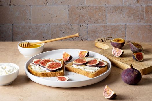 Bruschetta with figs, cream cheese and honey lie on light ceramic plate on light beige table against backdrop of masonry wall. Ingredients. Healthy food. Selective focus.