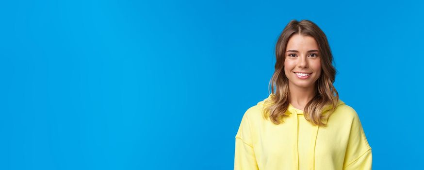 Close-up portrait gorgeous blond caucasian young girl with short haircut, wearing yellow hoodie, smiling with beaming white grin, looking camera, standing blue background. Copy space