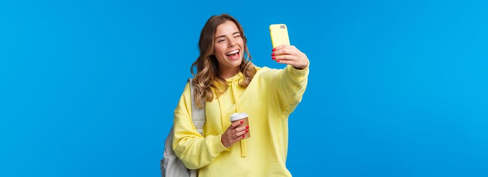 Lifestyle, university and education concept. Cheerful good-looking blond female student or traveller taking selfie with backpack and take-away coffee in paper cup, wink to mobile phone camera.