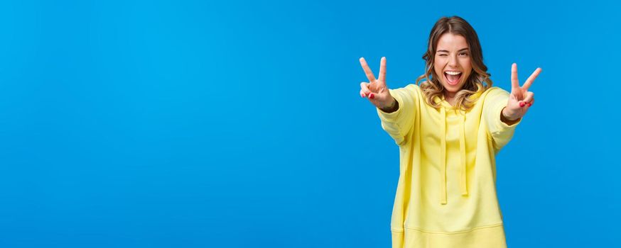 Stay positive. Kawaii joyful, friendly-looking smiling blond woman in yellow hoodie, stretch hands forward with peace gestures, show tongue and grinning, feel excited and upbeat.
