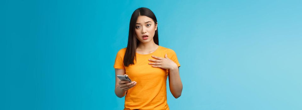 Offended shocked asian girl receive insulting message via internet social media, hold smartphone look surprised upset gasping, drop jaw touch chest shook, stand blue background. Copy space