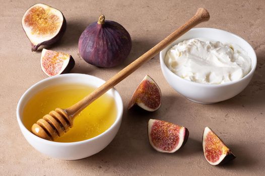 Cut fresh figs, honey in white bowl with wooden spoon and cream cheese on table. Healthy food. Selective focus.