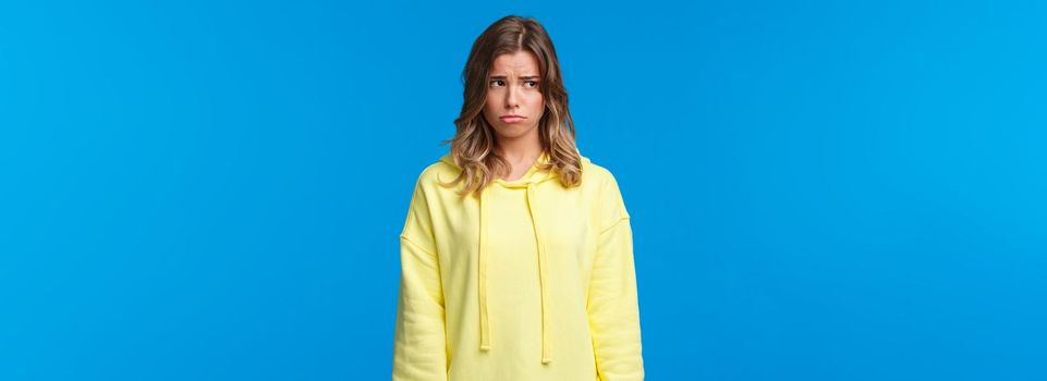 Gloomy and lonely say caucasian girl looking away, sobbing childish and sulking as feeling uneasy, being let down, having bad day at university, standing upset over blue background.