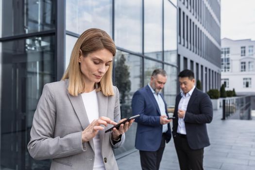 A successful and experienced business woman outside the office building is reading from a tablet computer, an employee with colleagues in business suits on a break