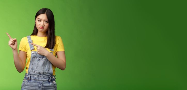 Disappointed uninterested asian gloomy girl pointing fingers left, grimacing sulking displeased, unwilling take part event, stare reluctant unimpressed, pose green background. Copy space