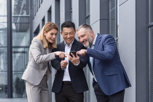 Diverse business group, three male and female workers outside office building cheerfully smiling and happy watching video on phone