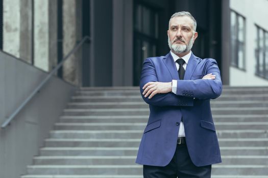 Portrait of a senior gray-haired man director, ceo on the background of the stairs of the office center. He is standing serious in a suit, arms crossed in front, looking to the side.