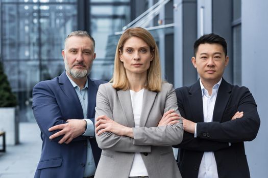 Successful and serious diverse team of three business people, man and woman focused looking at camera with arms crossed, portrait of co-workers outside office building