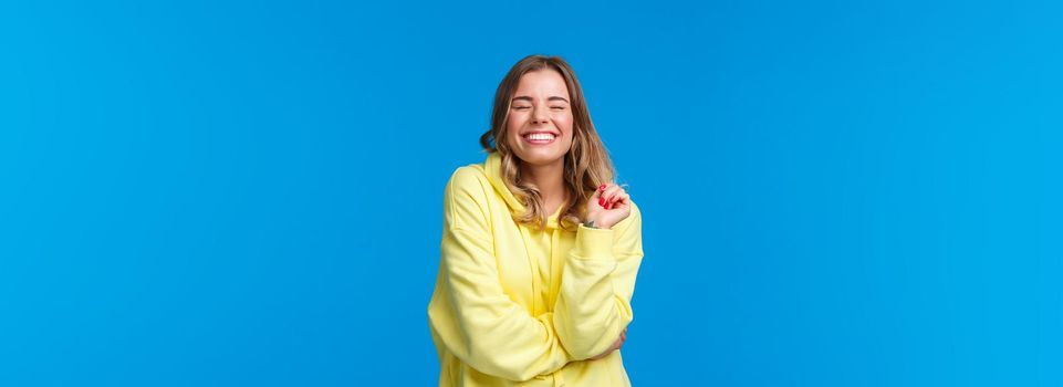 Cheerful carefree pleased girl feel happy and delighted, close eyes dreamy smiling with perfect beaming grin, touch strand of hair, standing in yellow hoodie over blue background.