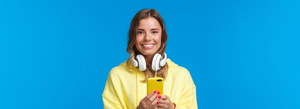 Close-up portrait of young blond european hipster girl with blond short hair and headphones over neck, holding smartphone, texting message, use mobile phone, blue background. Copy space