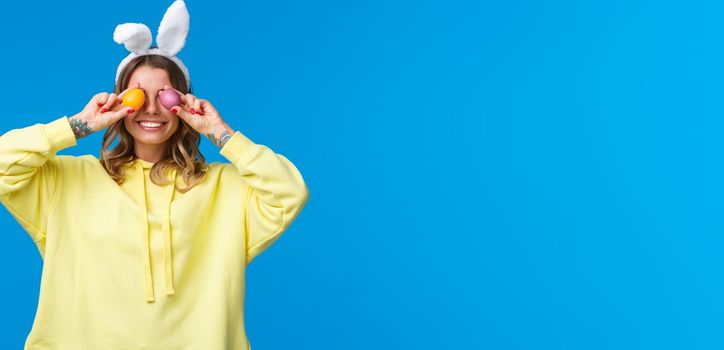 Holidays, traditions and celebration concept. Happy carefree smiling caucasian woman celebrating Easter, holding two painted eggs on eyes and grinning, wear cute rabbit ears, blue background.