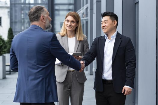 Meeting of three successful business people, diverse dream team man and woman outside office building, greeting and shaking hands, experienced professionals specialists in business suits talking