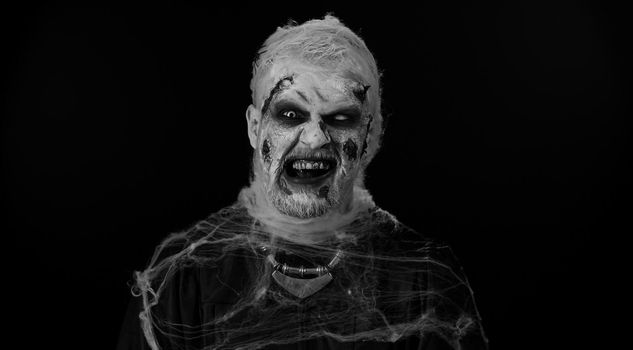 Zombie man with makeup with fake wounds scars and white contact lenses looking at camera and clicks his teeth, trying to scare in dark room. Sinister dead guy. Halloween, filming, staging concept
