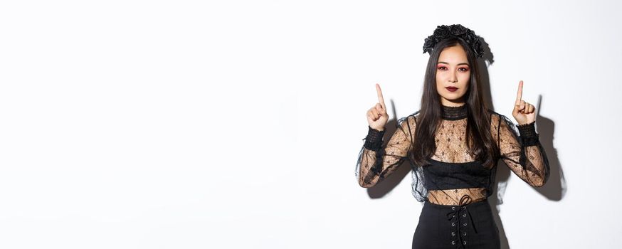 Sassy beautiful asian woman in black gothic dress, wearing witch costume for halloween and pointing fingers up, showing your logo or banner on empty white background, white background.