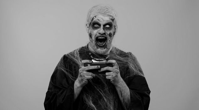 Spooky one man with horrible scary Halloween zombie makeup enthusiastically playing racing drive video games on mobile phone. Dead guy with wounded bloody scars face isolated against gray background