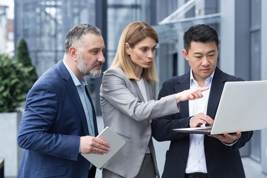 Successful business team, three colleagues businessman and businesswoman outside office building discussing current plans and management, looking at laptop screen, discussing and consulting