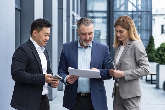 Diverse business team three colleagues employees discuss documents contract outside office building businessman and businesswoman holding bills and financial reports talking