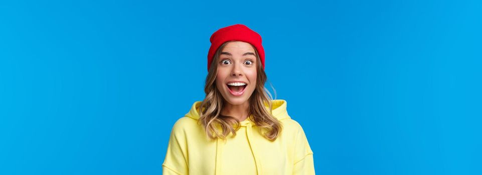 Close-up portrait of excited and intrigued young hipster girl in red beanie and yellow hoodie, gasping amazed look camera wondered and entertained, standing blue background.