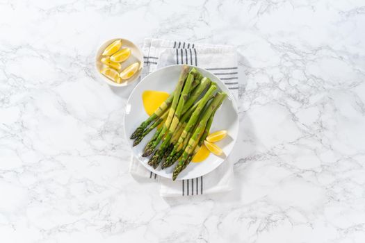 Flat lay. Serving steamed asparagus with homemade hollandaise sauce with lemon wedges on a white plate.