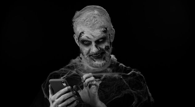 Frightening man with Halloween zombie bloody wounded makeup using mobile phone typing new post on web, sms message, browsing, addiction of social networks. Sinister undead guy on black wall background