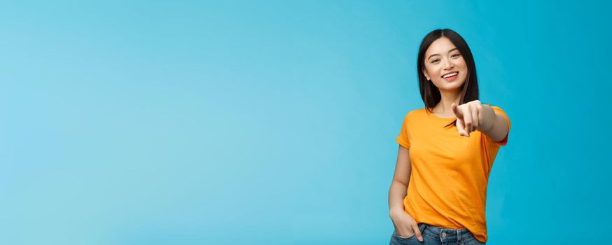 Lucky winner. Cheerful charismatic asian cute urban girl stand yellow t-shirt smiling friendly pointing finger camera choosing, picking person, inviting you team, stand happy blue background.