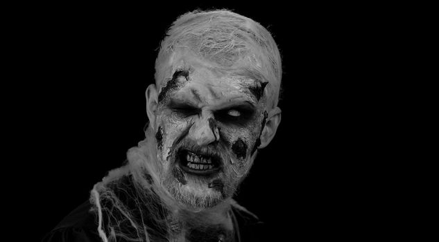 Zombie man with makeup with fake wounds scars and white contact lenses looking at camera and screaming, trying to scare in dark studio room. Sinister dead guy. Halloween, filming, staging concept