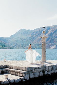 Bride in a flowing dress stands on a pier near a column against the backdrop of mountains. High quality photo