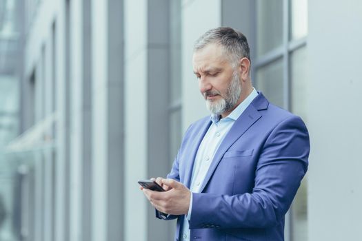 Close-up photo. Senior gray-haired man, a businessman in a suit, stands near the office center and calls a taxi from the phone, typing the number, waits, is in a hurry to meet.