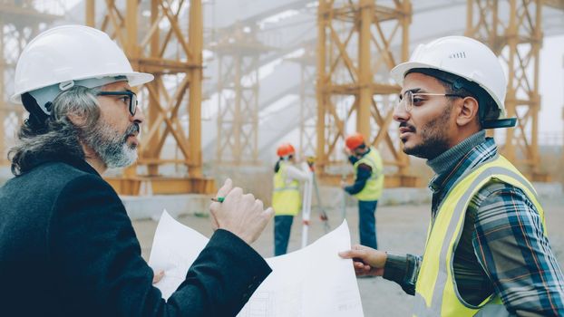 Foreman is discussing blueprints with project manager while surveyors are working in background. Construction site and workers concept.