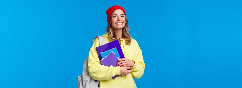 Charismatic smiling female in red beanie and yellow hoodie, walking to college, carry backpack and notebooks, papers for classes, look joyfully, studying prepare for exams to enter university.