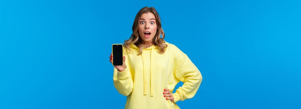 Impressed and surprised cute european girl showing amazing new app or mobile game, holding smartphone facing display to camera, staring speechless with excitement, blue background.