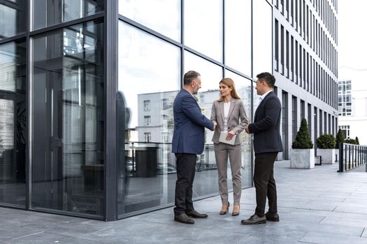 Meeting of three colleagues from outside the office building, experienced and mature IT specialists, greeting and shaking hands, business persons in business suits, diverse group of people