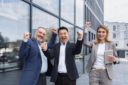 Successful diverse business team, three loving men and woman looking at camera and happy celebrating victory, team dreaming outside office building, business group in business suits