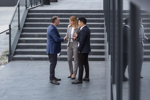 Meeting of three successful business people, diverse dream team man and woman outside office building, greeting and shaking hands, experienced professionals specialists in business suits talking