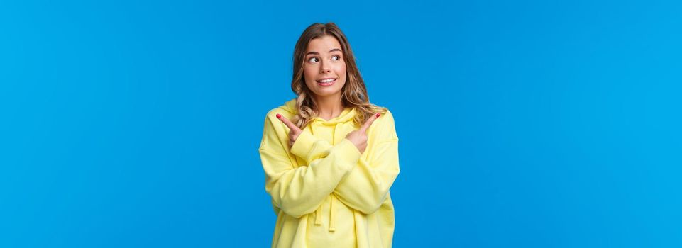 Various choices, cant decide. Cute dreamy blond girl in yellow hoodie making her choice, pointing fingers sideways at left and right side and smiling, picking one thing from all, blue background.