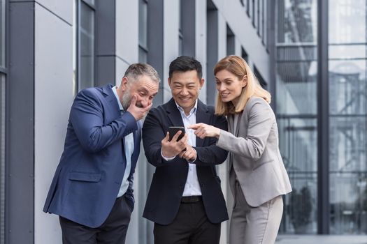 Diverse business group, three male and female workers outside office building cheerfully smiling and happy watching video on phone