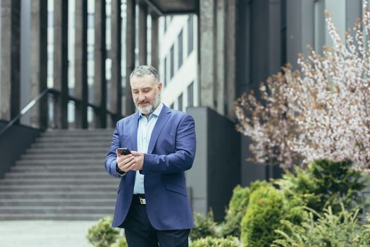 Senior gray-haired man, a businessman in a suit, stands near the modern office center and calls a taxi from the phone, typing the number, waits, is in a hurry to meet.