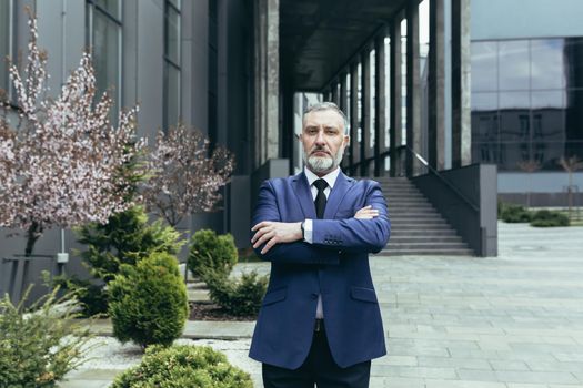 Portrait of senior handsome man, a businessman, a judge, a lawyer. Standing in a suit near the courthouse, office center. Crossed his arms in front, looks at the camera, serious.